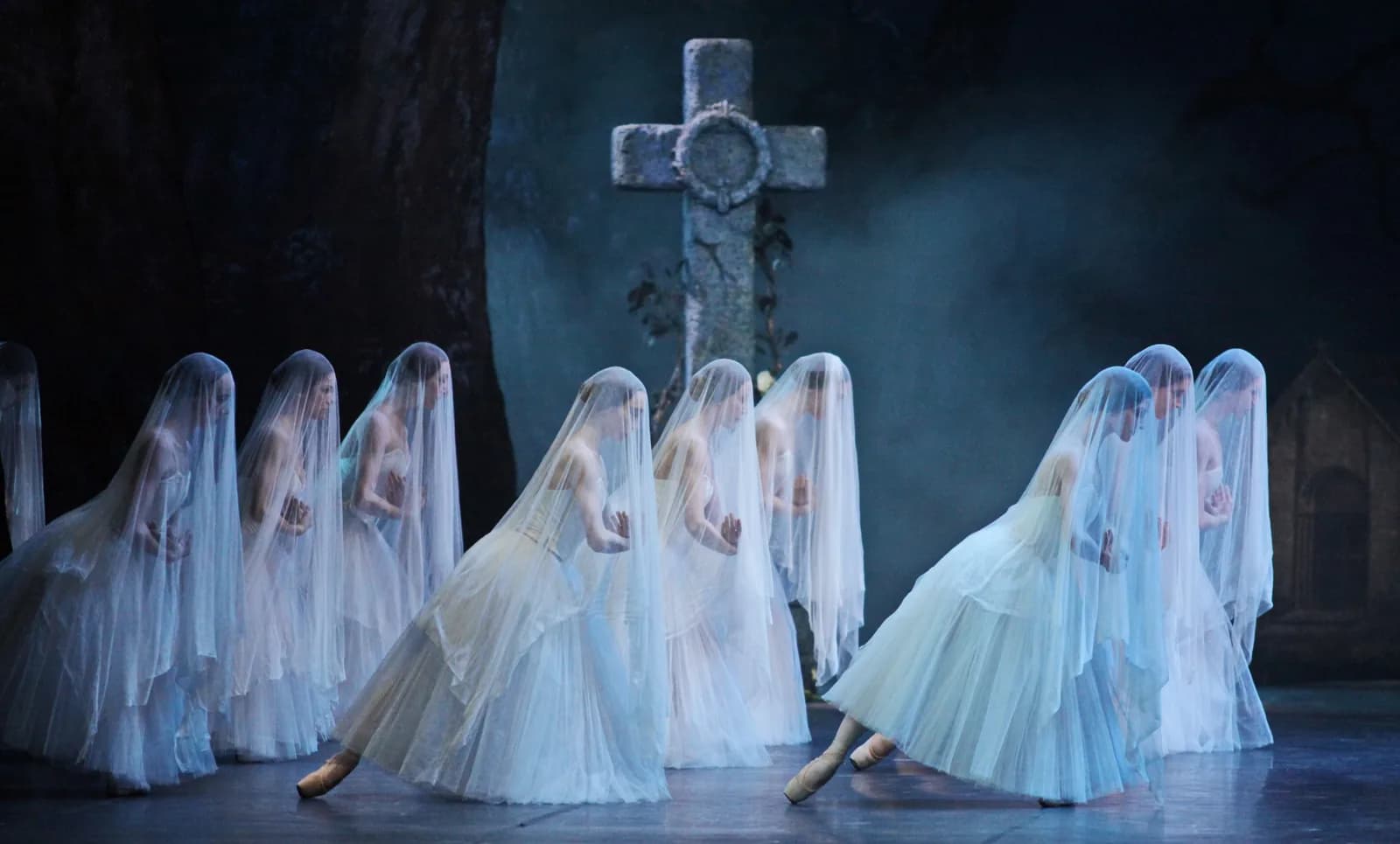 Des danseuses voilées de tulle groupées devant une croix de pierre