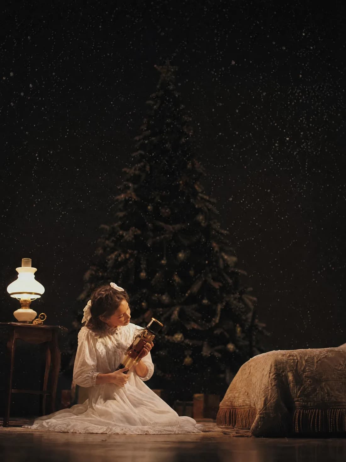 Une enfant en chemise de nuit tient un casse-noisette de bois près d'une lampe, un grand sapin sombre derrière