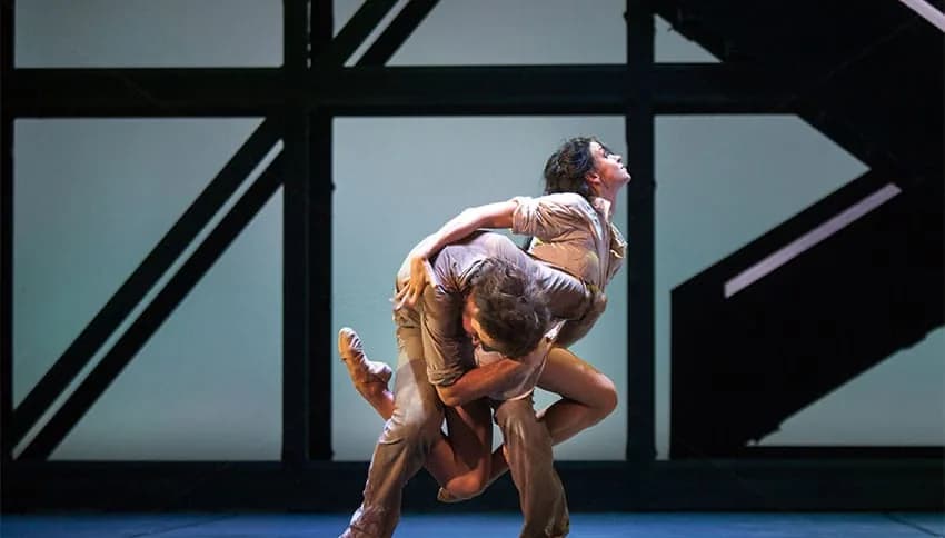 Deux danseurs en beige, l'un portant l'autre, devant une structure d'acier