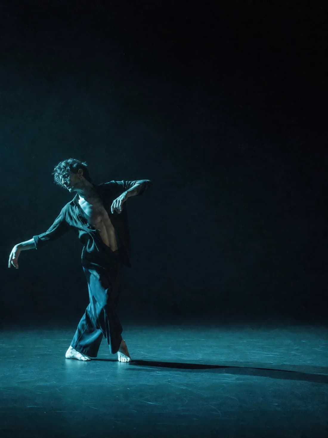 Un danseur seul sur un plateau vide, lumière latérale bleu-vert, longue ombre au sol