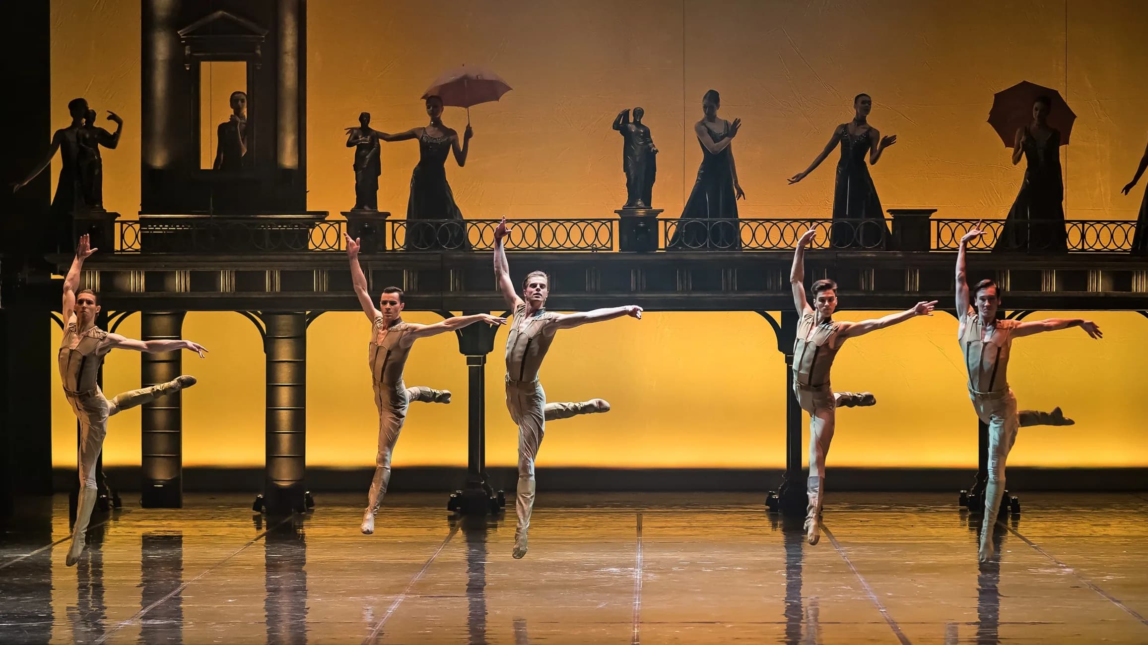 Cinq danseurs en combinaison claire sautent à l'unisson devant un fond sombre